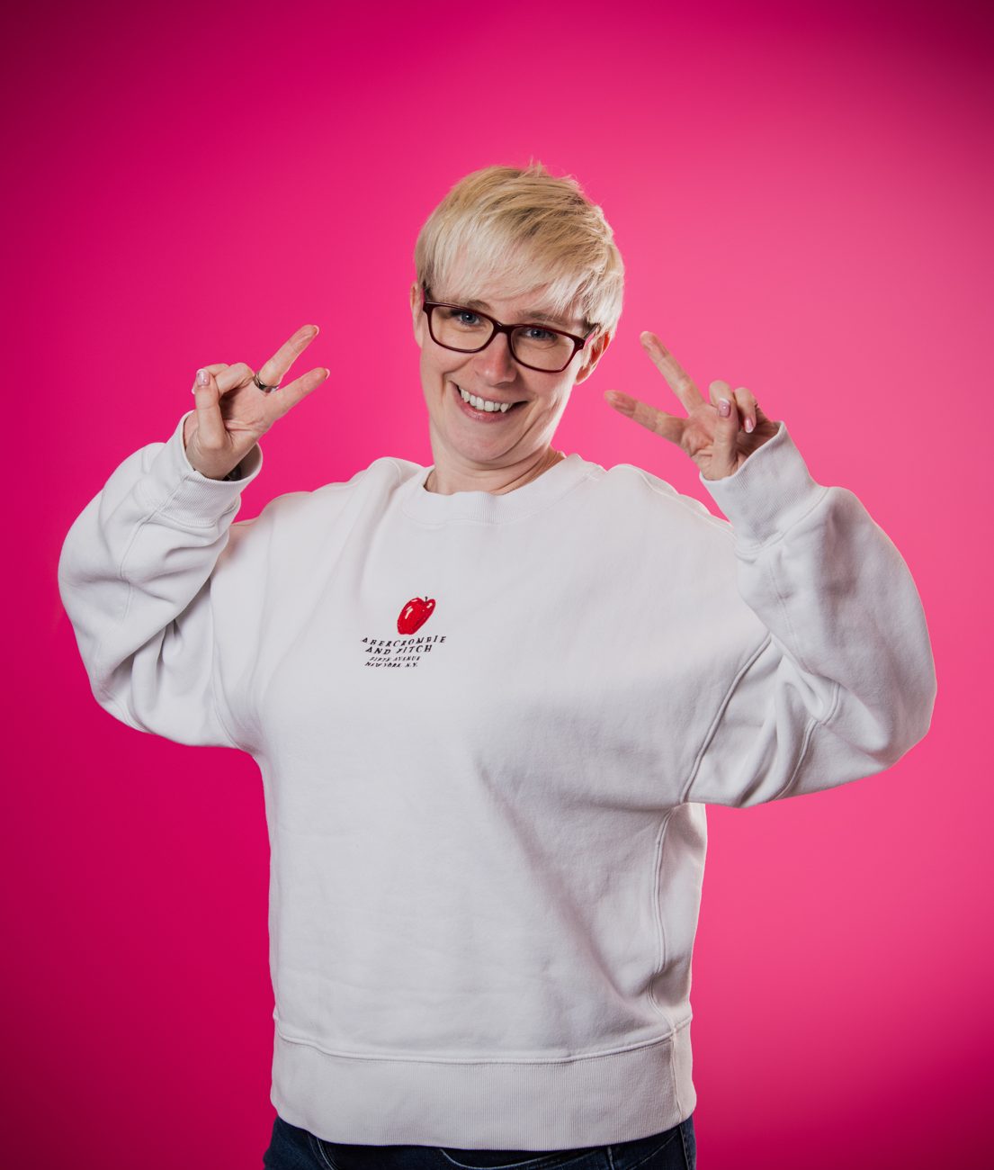 Jenny in a white sweatshirt against a bright pink wall, grinning and holding up two peace signs