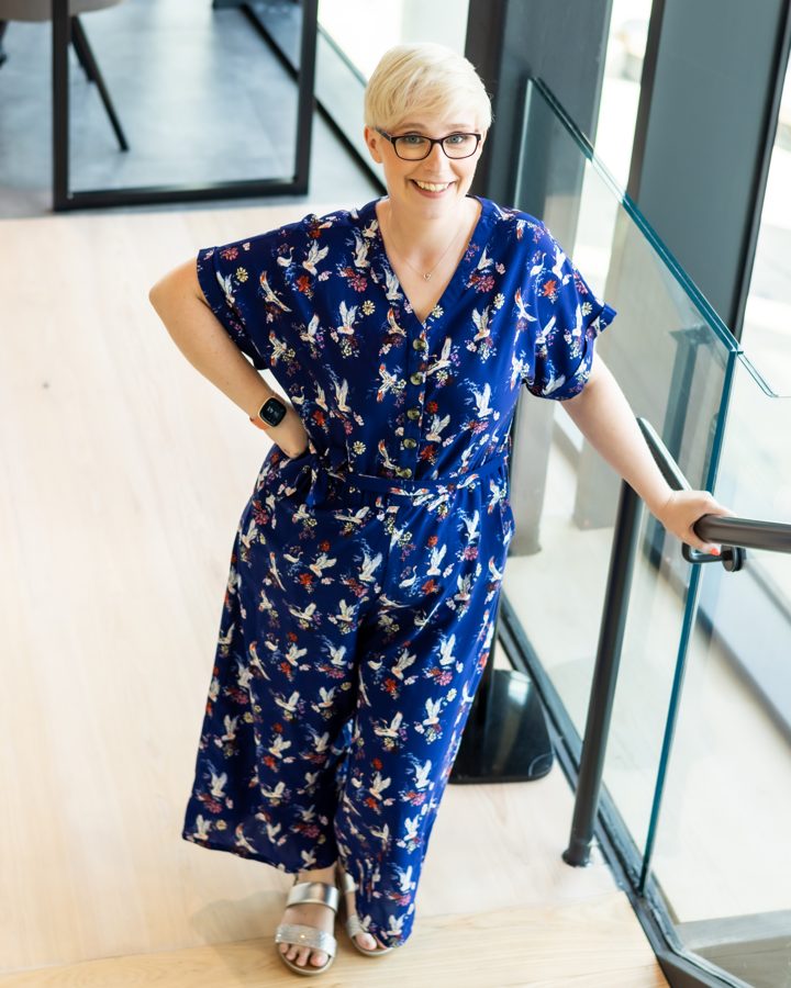 Jenny on a glass staircase in a blue patterned jumpsuit, hand on hip, smiling