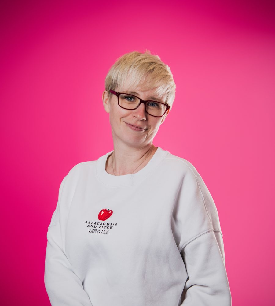 Jenny in a white sweatshirt against a bright pink wall, smiling with a raised eyebrow