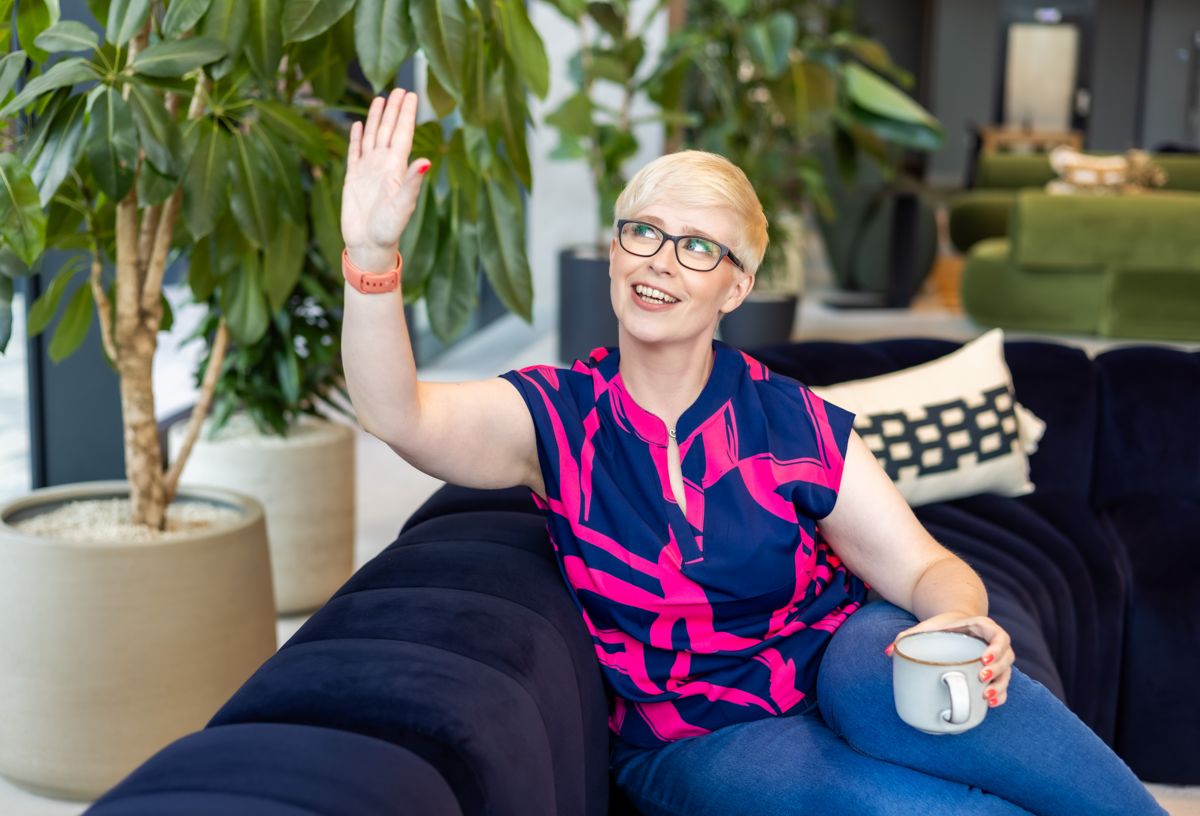 Jenny on a navy sofa, waving with a mug in her other hand